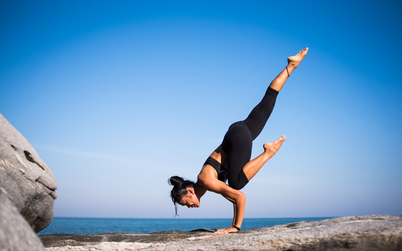 hero-img-01 Woman performing a yoga pose on a rocky beach with ocean view and clear blue sky.